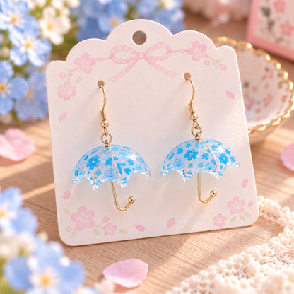 Paire de boucles d’oreilles fantaisie en forme de petits parapluies transparents, décorés de délicates fleurs bleues et de motifs floraux. Leurs fines attaches et manches légèrement dorés apportent une touche élégante et raffinée. Présentées sur un joli support rose pastel orné de fleurs de sakura, dans un décor romantique aux tons doux, ces boucles d’oreilles kawaii originales sont idéales pour compléter une tenue printanière, féminine et poétique.