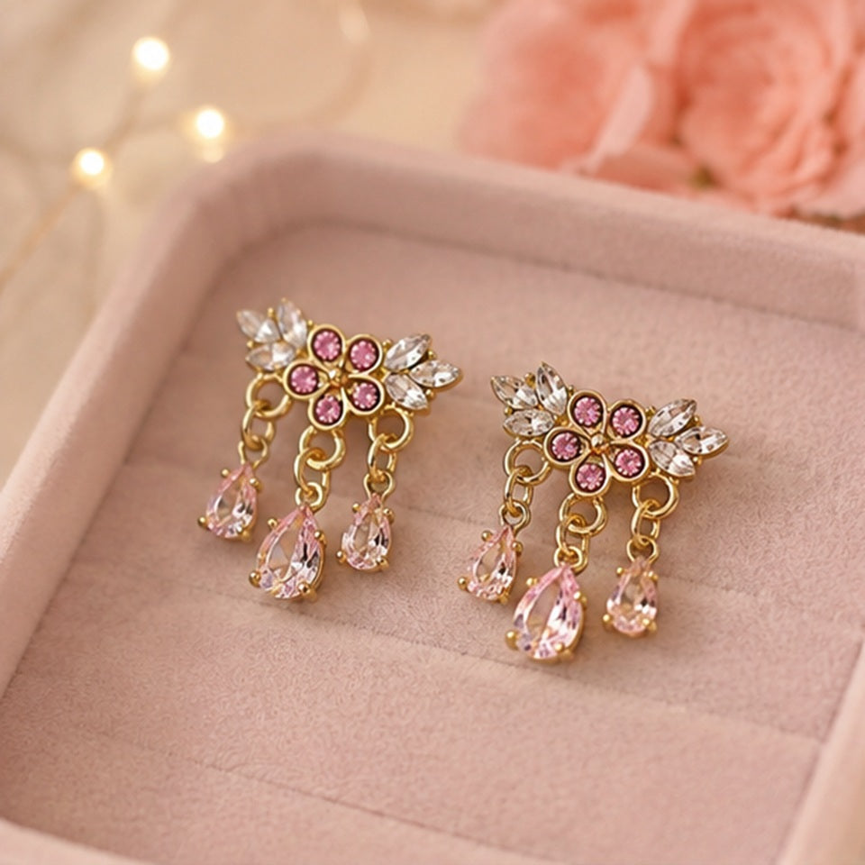 Des boucles d'oreilles florales dorées ornées de strass roses et blancs scintillants. Les bijoux sont présentés en gros plan dans une ambiance douce et féminine, mettant en valeur leurs pampilles en cristal rose et leur éclat lumineux.