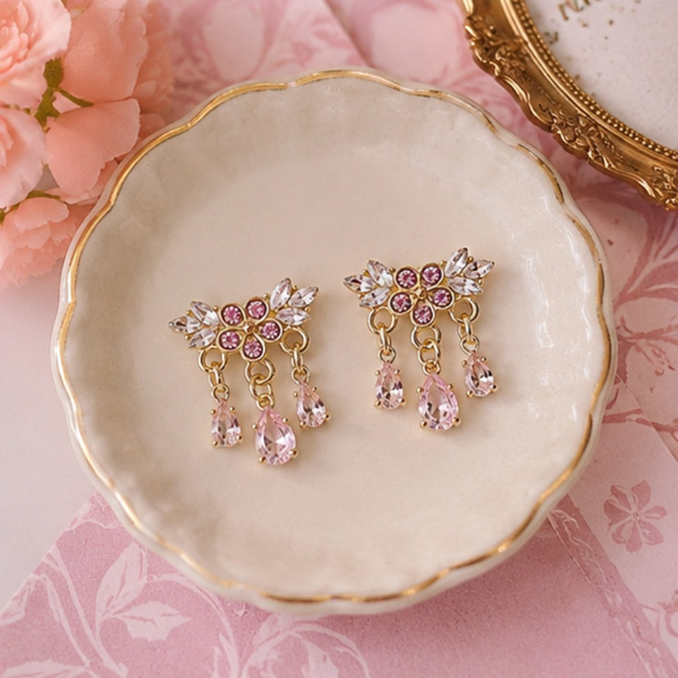 Des boucles d'oreilles florales dorées ornées de strass roses et blancs scintillants. Les bijoux sont présentés en gros plan dans une ambiance douce et féminine, mettant en valeur leurs pampilles en cristal rose et leur éclat lumineux.