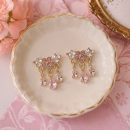 Des boucles d'oreilles florales dorées ornées de strass roses et blancs scintillants. Les bijoux sont présentés en gros plan dans une ambiance douce et féminine, mettant en valeur leurs pampilles en cristal rose et leur éclat lumineux.