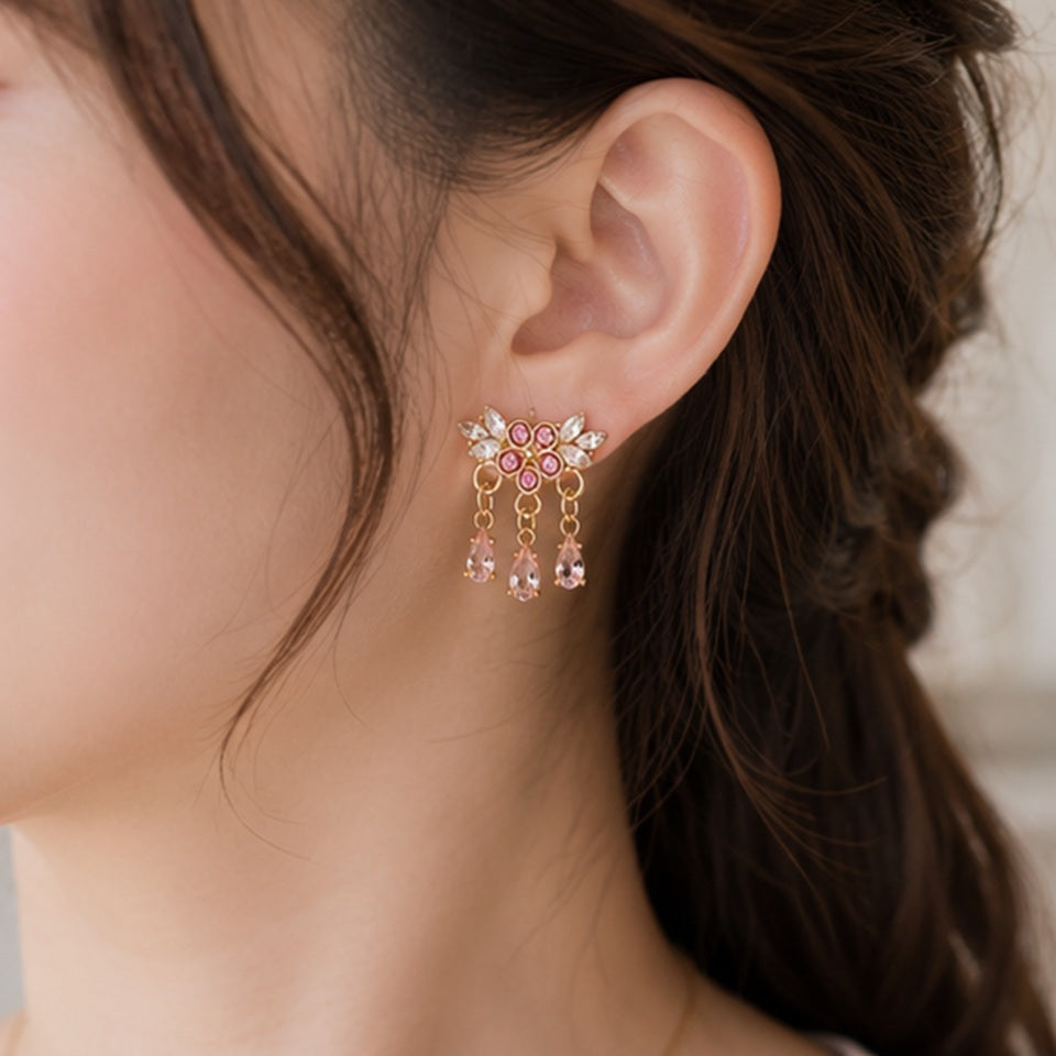 Des boucles d'oreilles florales dorées ornées de strass roses et blancs scintillants. Les bijoux sont présentés en gros plan dans une ambiance douce et féminine, mettant en valeur leurs pampilles en cristal rose et leur éclat lumineux.