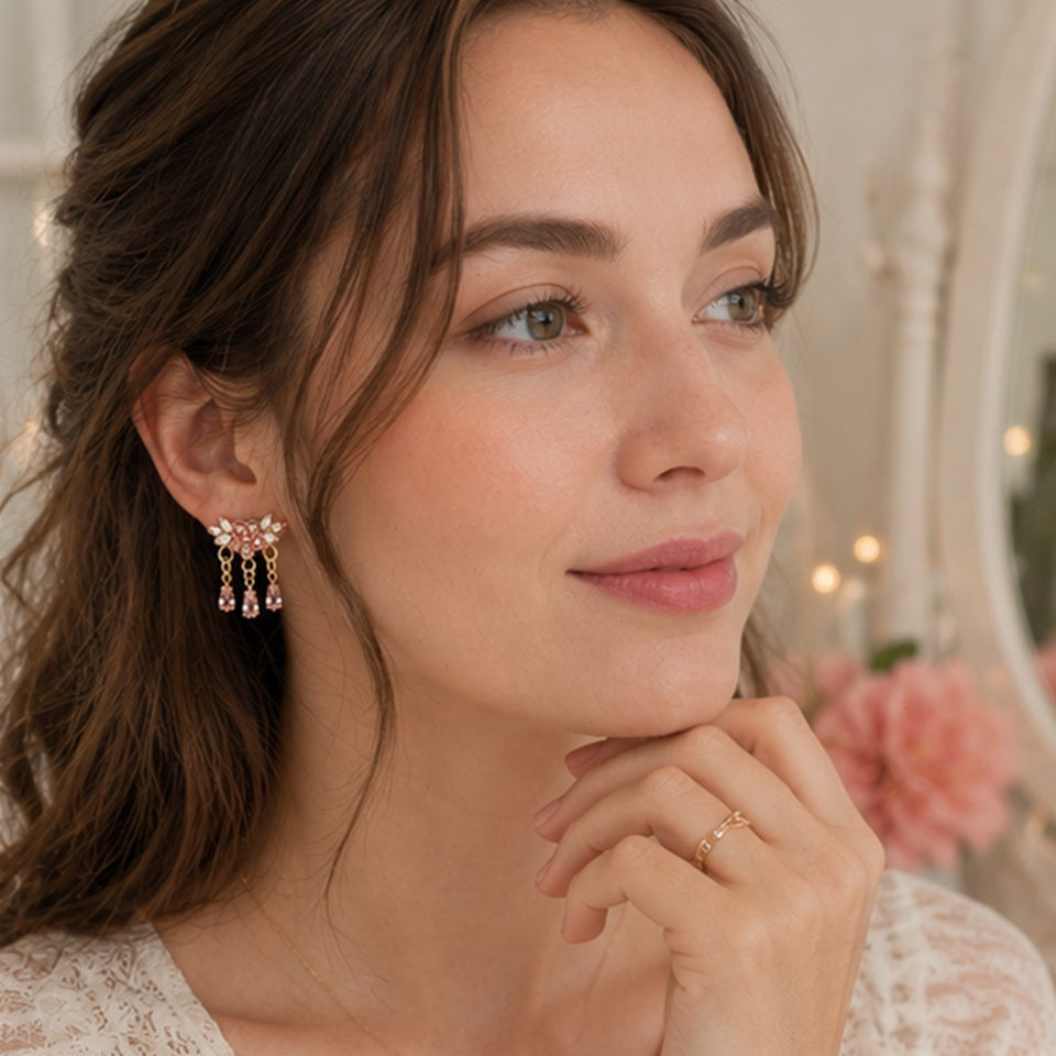 Des boucles d'oreilles florales dorées ornées de strass roses et blancs scintillants. Les bijoux sont présentés en gros plan dans une ambiance douce et féminine, mettant en valeur leurs pampilles en cristal rose et leur éclat lumineux.