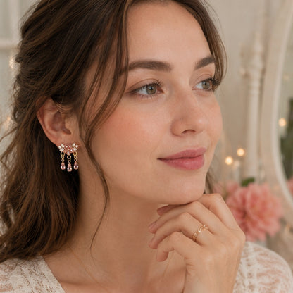 Des boucles d'oreilles florales dorées ornées de strass roses et blancs scintillants. Les bijoux sont présentés en gros plan dans une ambiance douce et féminine, mettant en valeur leurs pampilles en cristal rose et leur éclat lumineux.