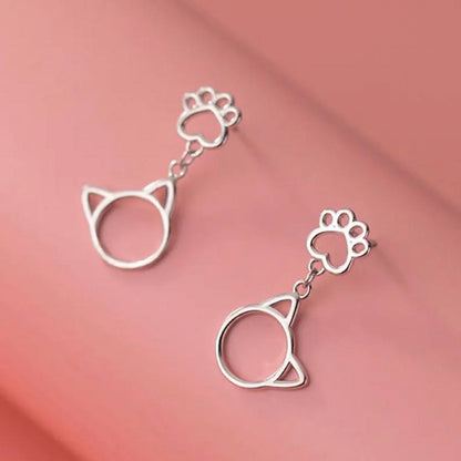 une paire de boucles d'oreilles argentée en forme de patte de chat d'où pendent de mignones breloques en forme de tête de chat.