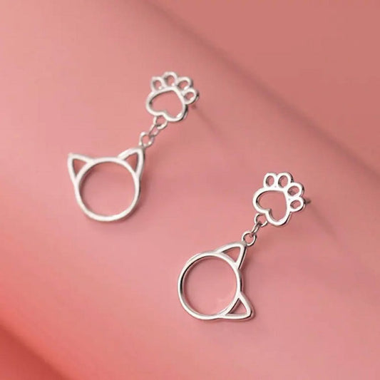 une paire de boucles d'oreilles argentée en forme de patte de chat d'où pendent de mignones breloques en forme de tête de chat.