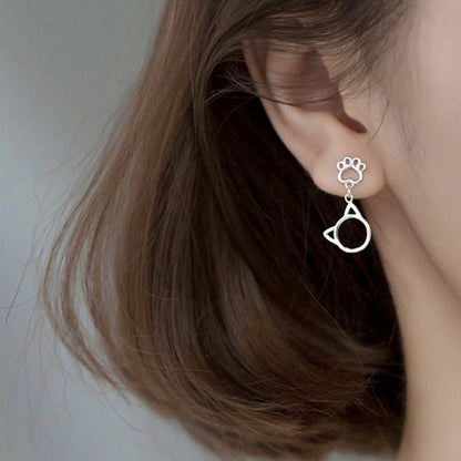 une paire de boucles d'oreilles argentée en forme de patte de chat d'où pendent de mignones breloques en forme de tête de chat.
