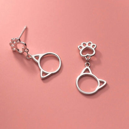 une paire de boucles d'oreilles argentée en forme de patte de chat d'où pendent de mignones breloques en forme de tête de chat.