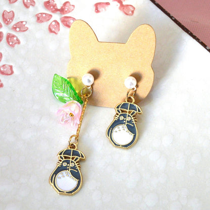 Une paire de boucles d'oreilles asymétriques Totoro qui porte son parapluie, de l'autre côté se trouve une jolie fleur rose et une feuille verte