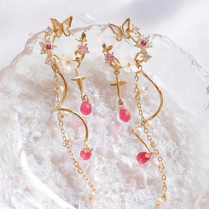 une paire de boucles d'oreilles composées d'une ronde de fleurs blanches et de strass roses, d'un joli papillon blanc, d'où pendent une chaine dorée et une tige torsadée ainsi qu'une douce étoilée dorée. Au bout, il y a des perles en forme de goutte rose. l'ensemble est poétique et féminin.