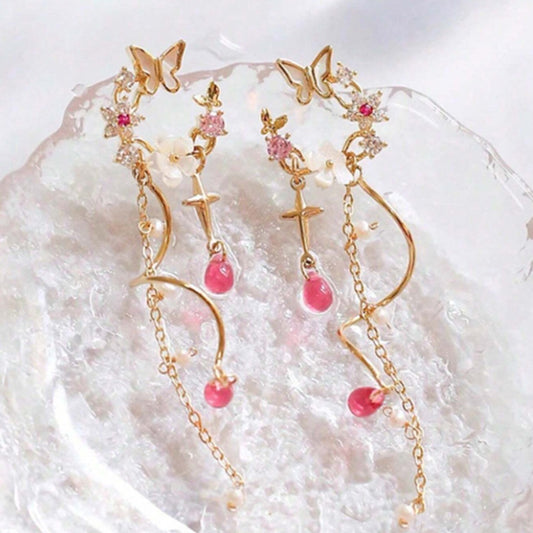 une paire de boucles d'oreilles composées d'une ronde de fleurs blanches et de strass roses, d'un joli papillon blanc, d'où pendent une chaine dorée et une tige torsadée ainsi qu'une douce étoilée dorée. Au bout, il y a des perles en forme de goutte rose. l'ensemble est poétique et féminin.