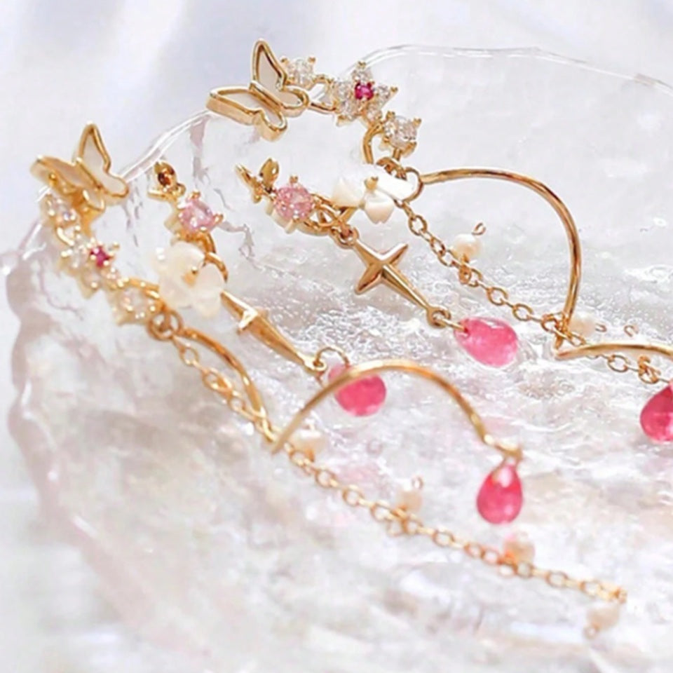une paire de boucles d'oreilles composées d'une ronde de fleurs blanches et de strass roses, d'un joli papillon blanc, d'où pendent une chaine dorée et une tige torsadée ainsi qu'une douce étoilée dorée. Au bout, il y a des perles en forme de goutte rose. l'ensemble est poétique et féminin.