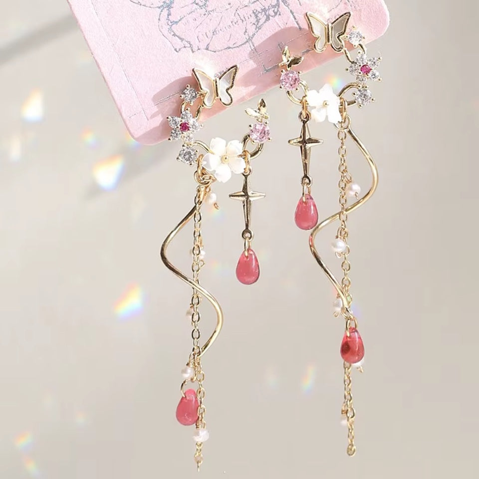 une paire de boucles d'oreilles composées d'une ronde de fleurs blanches et de strass roses, d'un joli papillon blanc, d'où pendent une chaine dorée et une tige torsadée ainsi qu'une douce étoilée dorée. Au bout, il y a des perles en forme de goutte rose. l'ensemble est poétique et féminin.