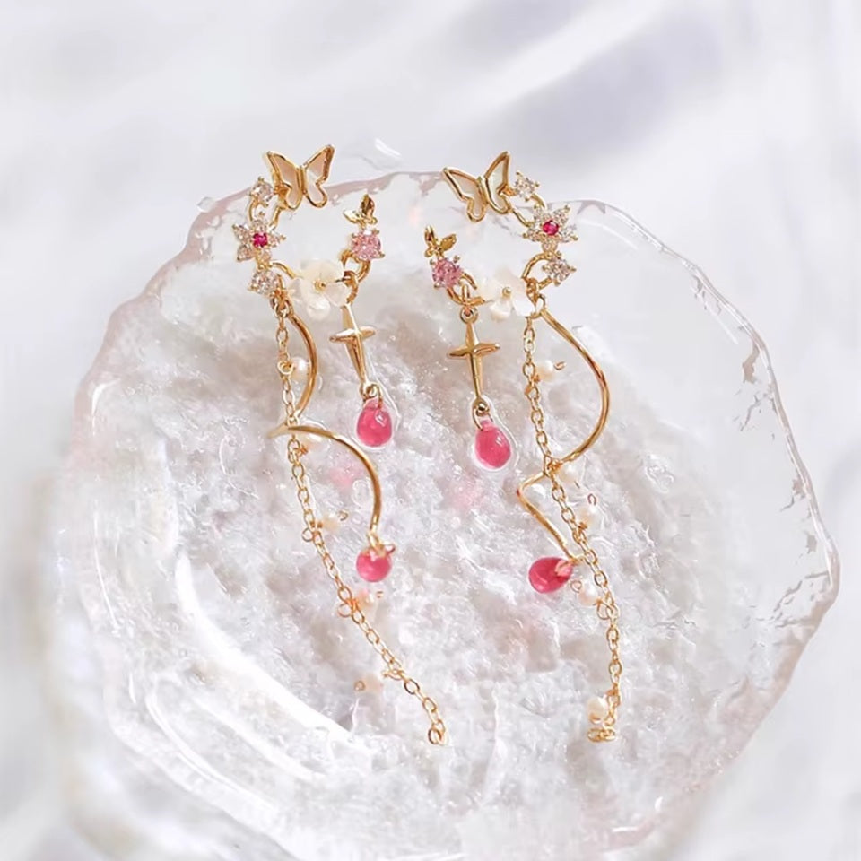 une paire de boucles d'oreilles composées d'une ronde de fleurs blanches et de strass roses, d'un joli papillon blanc, d'où pendent une chaine dorée et une tige torsadée ainsi qu'une douce étoilée dorée. Au bout, il y a des perles en forme de goutte rose. l'ensemble est poétique et féminin.