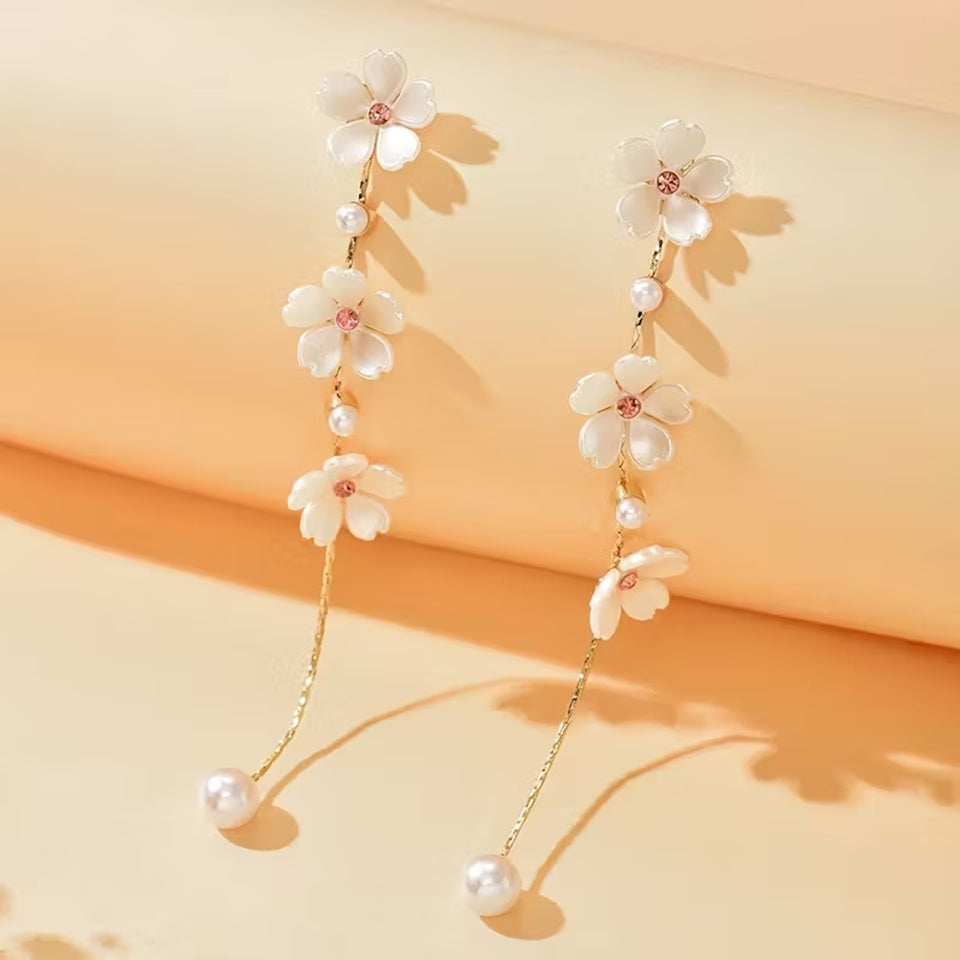 une superbe paire de boucles d'oreilles présentant trois fleurs de sakura blanches dont le coeur est un strass rose suspendues à une chaine dorée qui termine par une perle blanche. Le bijou est élégant, féminin et poétique.