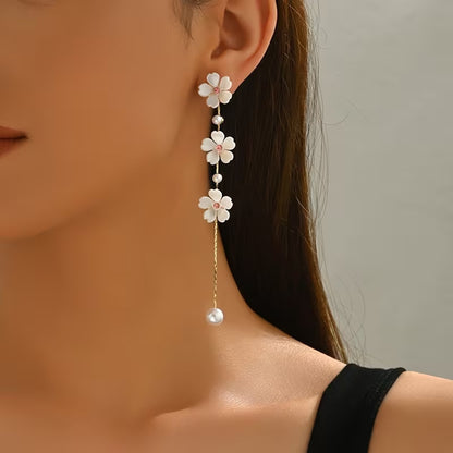 une superbe paire de boucles d'oreilles présentant trois fleurs de sakura blanches dont le coeur est un strass rose suspendues à une chaine dorée qui termine par une perle blanche. Le bijou est élégant, féminin et poétique.