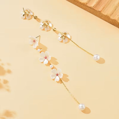 une superbe paire de boucles d'oreilles présentant trois fleurs de sakura blanches dont le coeur est un strass rose suspendues à une chaine dorée qui termine par une perle blanche. Le bijou est élégant, féminin et poétique.