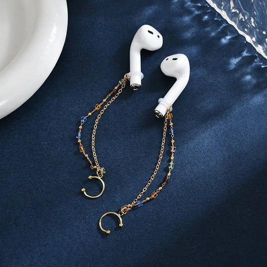 des boucles décoratives composées de pierres de couleur bleu, orange et verte attachées à des écouteurs sans fils blancs et posés sur une table bleue. 