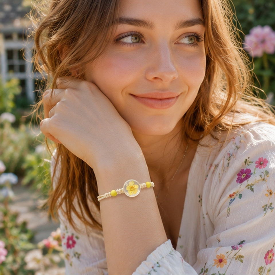 Portrait d'une femme dans un jardin fleuri portant un bracelet à perles jaunes et cabochon transparent contenant une fleur de prunier séchée. Le bracelet est mis en avant grâce à une pose élégante, tandis que les massifs de fleurs colorées offrent un décor lumineux, doux et bohème.
