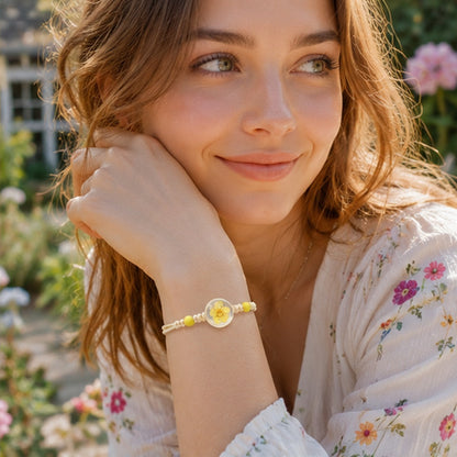 Portrait d'une femme dans un jardin fleuri portant un bracelet à perles jaunes et cabochon transparent contenant une fleur de prunier séchée. Le bracelet est mis en avant grâce à une pose élégante, tandis que les massifs de fleurs colorées offrent un décor lumineux, doux et bohème.