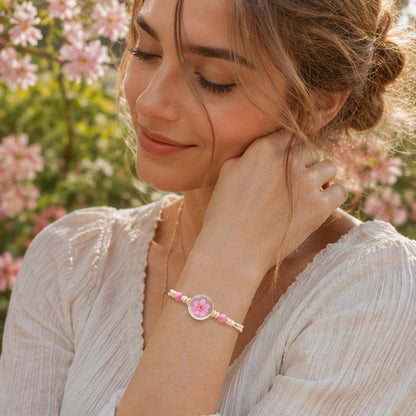 Femme portant un bracelet en corde beige orné d'un cabochon transparent renfermant une fleur de prunier rose séchée. Elle est vêtue d'un chemisier blanc et pose dans un jardin fleuri baigné de lumière naturelle. Les fleurs roses en arrière-plan créent une ambiance romantique et printanière qui met en valeur le bijou.