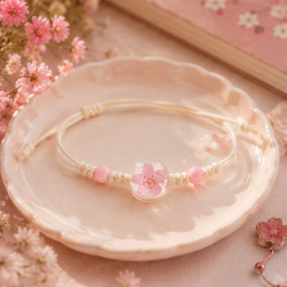 Bracelet en corde beige orné d'un cabochon transparent renfermant une fleur de prunier séchée rose et de perles assorties. Il est présenté sur une coupelle en céramique blanche, entouré de petites fleurs roses et d'accessoires décoratifs dans une ambiance douce, romantique et printanière.