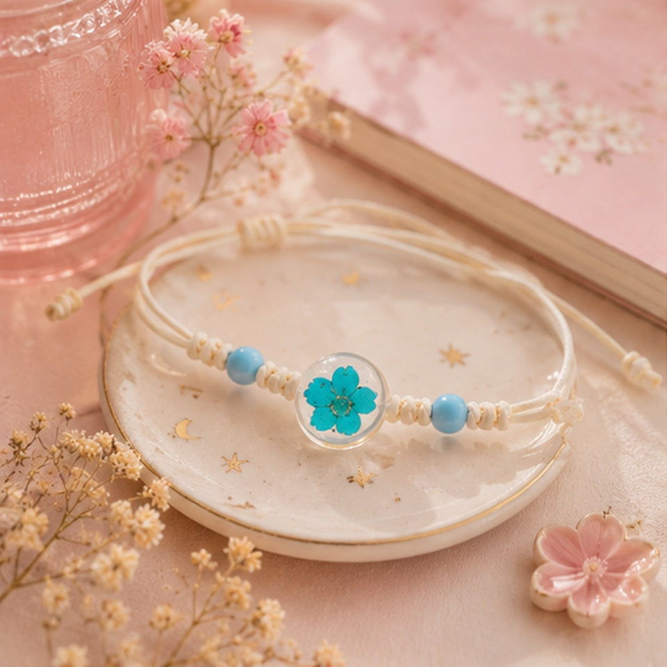 Bracelet en corde beige agrémenté de perles bleues et d'un cabochon transparent mettant en valeur une fleur de prunier séchée bleue. Présenté sur une petite assiette en céramique, il est accompagné de fleurs séchées, d'un carnet fleuri et d'une verrerie rose pour une ambiance bohème élégante.