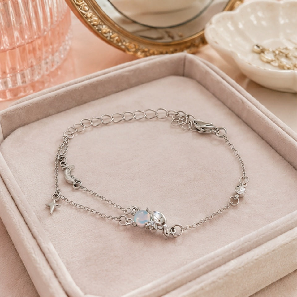un bracelet argenté représentant Cinnamoroll tenant une pierre de lune. Il a deux chaines ornées d'une étoile et d'une lune. Il est posé dans une jolie boite en velours sur une coiffeuse féminine.