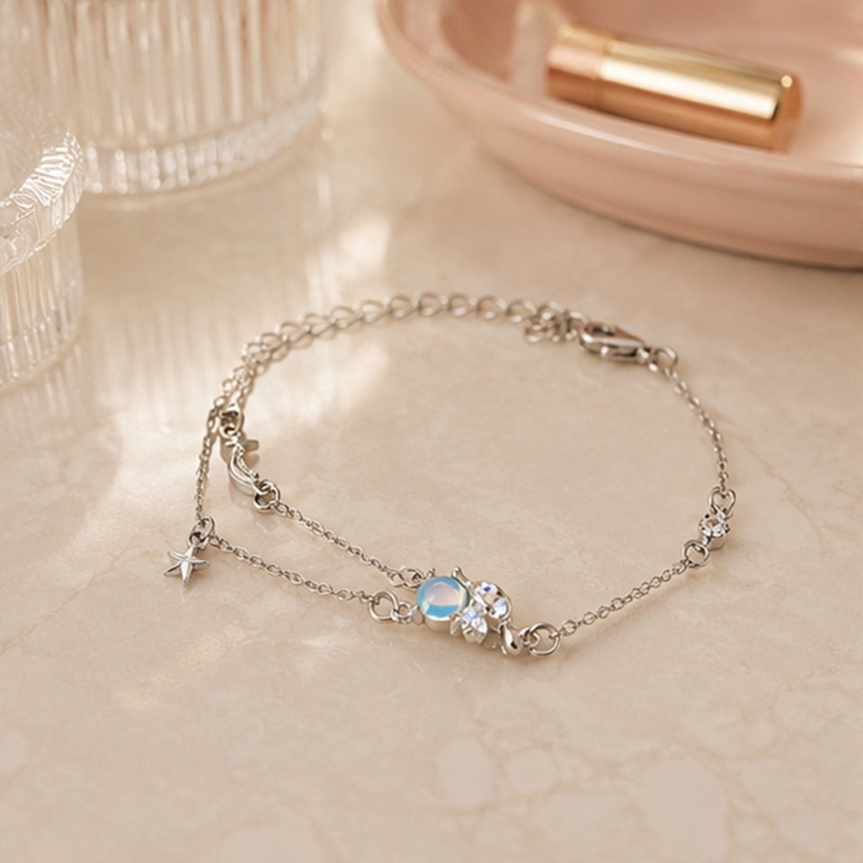un bracelet argenté représentant Cinnamoroll tenant une pierre de lune. Il a deux chaines ornées d'une étoile et d'une lune. Il est posé sur une coiffeuse féminine dans une ambiance douce.