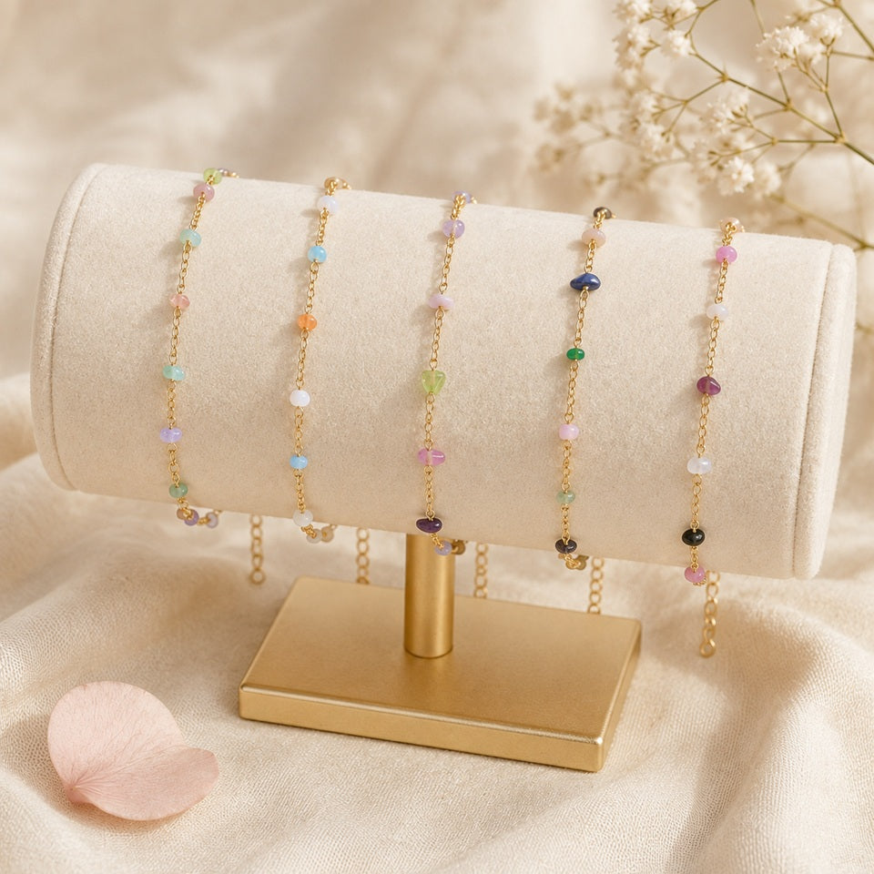 Des bracelets dorés fins ornés de pierres colorées, présentés sur un support cylindrique en velours beige avec une base dorée.