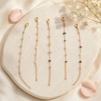 Des bracelets dorés fins ornés de petites pierres colorées, présentés sur un plateau en pierre claire avec des fleurs blanches et un pétale rose.