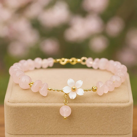 un bracelet doré composé de pierres et de perles roses pâles et d'une fleur de sakura. L'ensemble est raffiné et élégant.