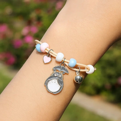 un bracelet en corde avec une breloque Totoro qui tient un parapluie. Sur le bracelet, il ya des perles bleu, rose et blanches avec un joli coeur rose et une clochette.