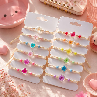 une collection de bracelets en corde ornés de véritables fleurs séchées sous cabochon transparent. Les bracelets, aux perles pastel de différentes couleurs, sont disposés sur des cartons de présentation entièrement blancs, sans aucun texte. L'ensemble est mis en scène dans un décor féminin et délicat, le tout baigné par une lumière naturelle douce qui met en valeur les détails floraux et les couleurs printanières