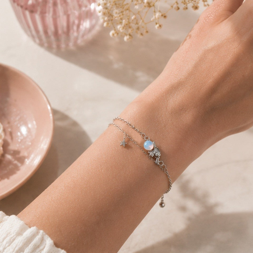 un bracelet argenté représentant Cinnamoroll tenant une pierre de lune. Il a deux chaines ornées d'une étoile et d'une lune. Le décor est dans les tons pastels, roses dans une ambiance douce et calme.