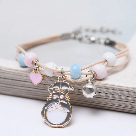 un bracelet en corde avec une breloque Totoro qui tient un parapluie. Sur le bracelet, il ya des perles bleu, rose et blanches avec un joli coeur rose et une clochette.