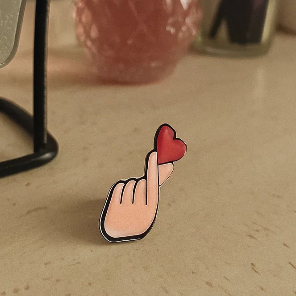 une broche en forme d'une main qui forme un finger heart, ce coeur coréen symbole d'amour. Au bout des doigts se trouve un coeur rouge