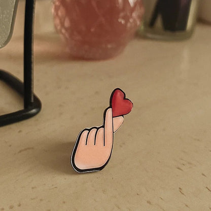 une broche en forme d'une main qui forme un finger heart, ce coeur coréen symbole d'amour. Au bout des doigts se trouve un coeur rouge