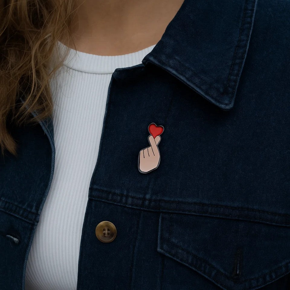 une broche en forme d'une main qui forme un finger heart, ce coeur coréen symbole d'amour. Au bout des doigts se trouve un coeur rouge