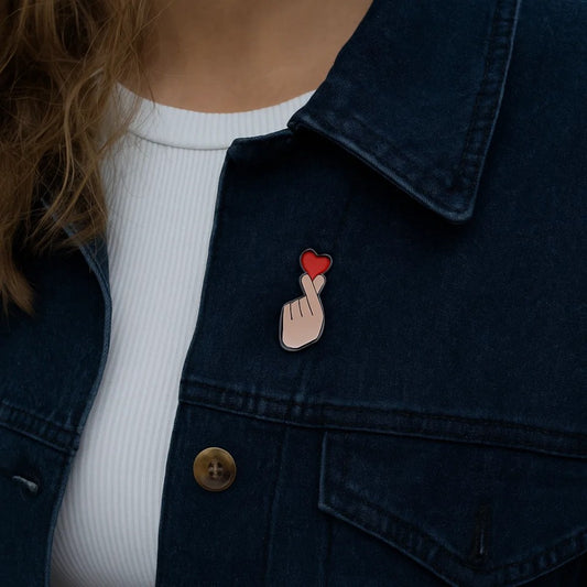 une broche en forme d'une main qui forme un finger heart, ce coeur coréen symbole d'amour. Au bout des doigts se trouve un coeur rouge