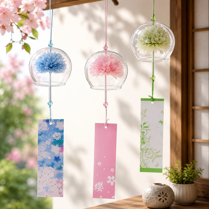 trois carillons japonais suspendus dans une véranda lumineuse. Chaque carillon est constitué d'une clochette en verre transparent renfermant une fleur délicate : une fleur bleue à gauche, une rose au centre et une vert clair à droite. De longues étiquettes décoratives aux motifs floraux pendent sous chaque cloche. L'ensemble est suspendu à une poutre en bois, avec des fleurs de cerisier floues en arrière-plan et une ambiance douce, printanière et apaisante.