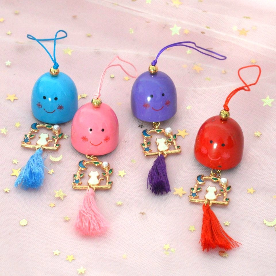 un carillon avec une clochette expression kawaii, une breloque en forme de chat qui est sur une balançoire y est suspendu, et un pompon coloré finit l'ensemble.