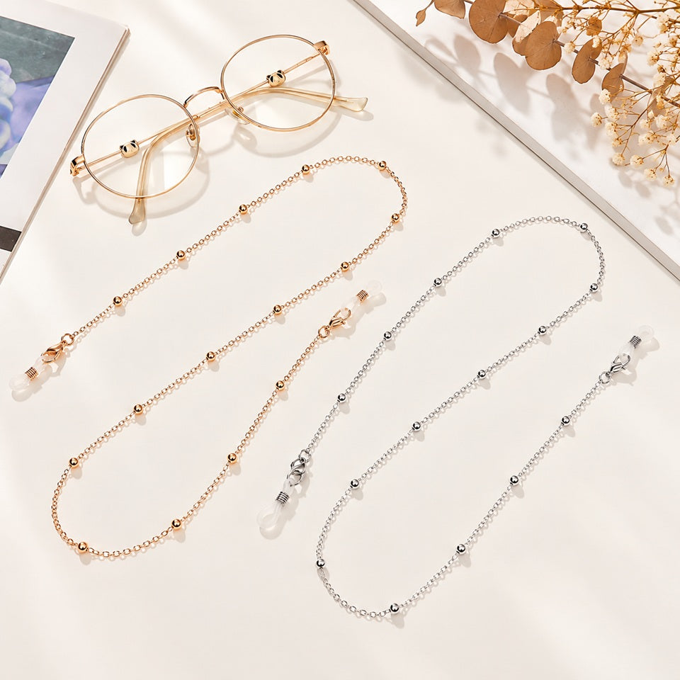 Chaînes à lunettes chic et élégantes, dorée rose et argentée, ornées de petites boules métalliques, avec embouts en silicone, présentées à côté de lunettes rondes sur fond clair. Accessoires de mode raffinés et originaux.