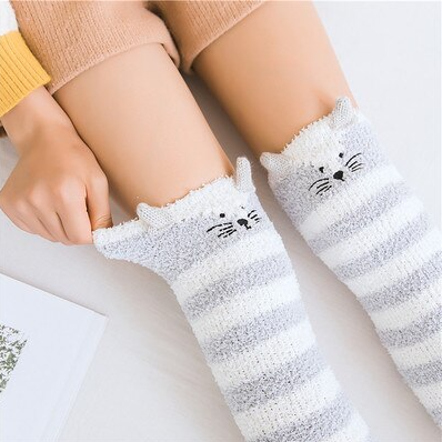 une paire de chaussettes hautes rayées grises et blanches en matière pilou toutes douces. Sur le haut des chaussettes est brodé un visage de chat avec ses moustaches et deux petites oreilles