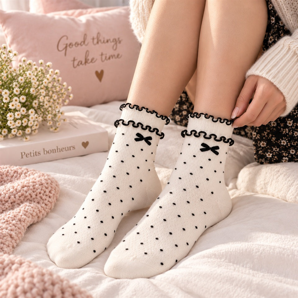 Femme portant des chaussettes blanches à pois noirs avec petits nœuds et double froufrou, sans chaussures, dans une ambiance douce et féminine.