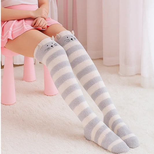 une paire de chaussettes hautes rayées grises et blanches en matière pilou toutes douces. Sur le haut des chaussettes est brodé un visage de chat avec ses moustaches et deux petites oreilles