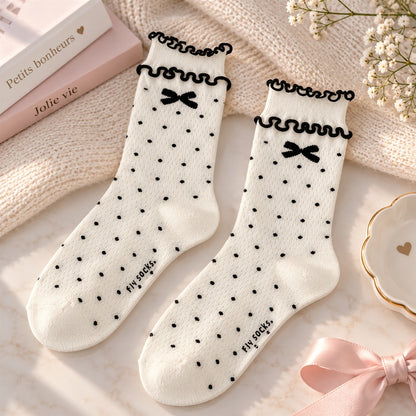 Chaussettes blanches à pois noirs avec petits nœuds et double bord à froufrous, présentées sur un fond doux et élégant aux tons crème et rose.