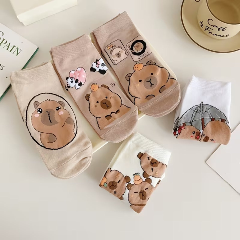 des chaussettes marrons et blanches au motif d'adorables capybaras posées et pliées sur une table blanche. Elles sont toutes douces et confortables. Les capybaras ont des expressions adorables et mignonnes.