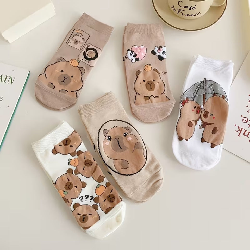 des chaussettes marrons et blanches au motif d'adorables capybaras posées sur une table blanche. Elles sont toutes douces et confortables. Les capybaras ont des expressions adorables et mignonnes.