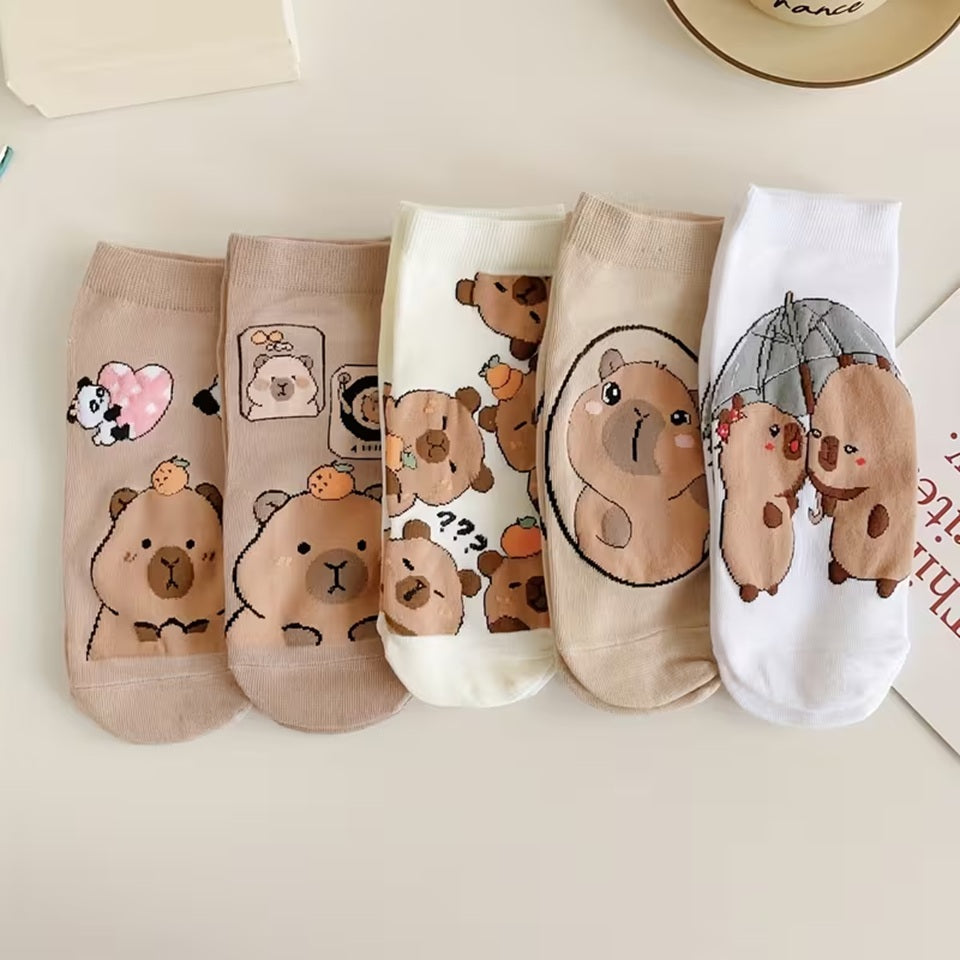 des chaussettes marrons et blanches au motif d'adorables capybaras posées et alignées sur une table blanche. Elles sont toutes douces et confortables. Les capybaras ont des expressions adorables et mignonnes.
