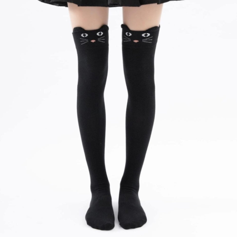 une paire de chaussettes hautes noires sur lesquelles est brodé un visage de chat avec ses moustaches. Elles ont deux petites oreilles qui dépassent du haut de la chaussette